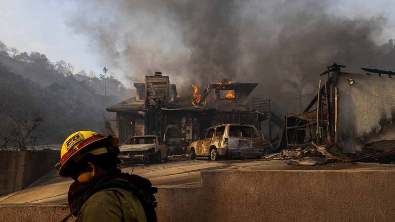 Firefighter in the foreground walks by a burning house in the background.